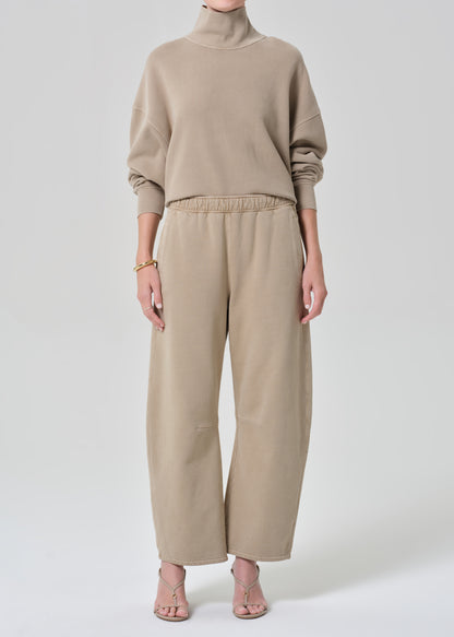Person wearing a beige turtleneck sweater and pants on a grey background