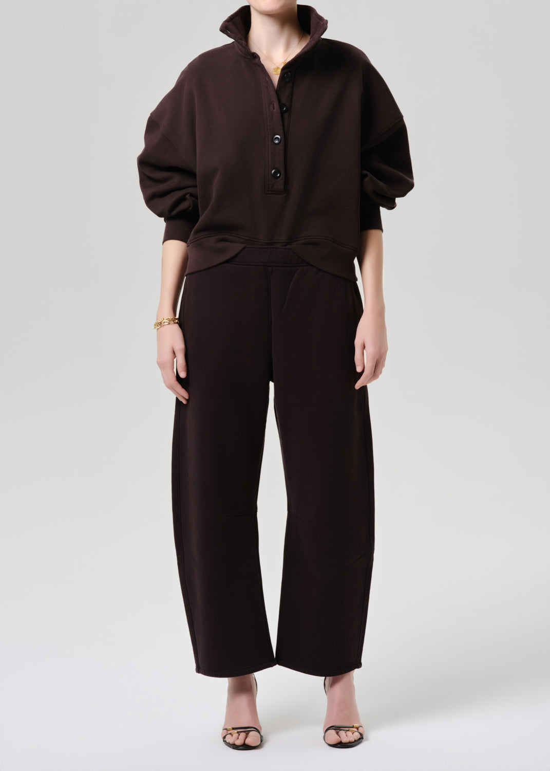 A model wearing a dark brown two-piece set consisting of a long-sleeve buttoned top and matching wide-leg cropped pants, paired with clear high-heeled sandals against a grey background.