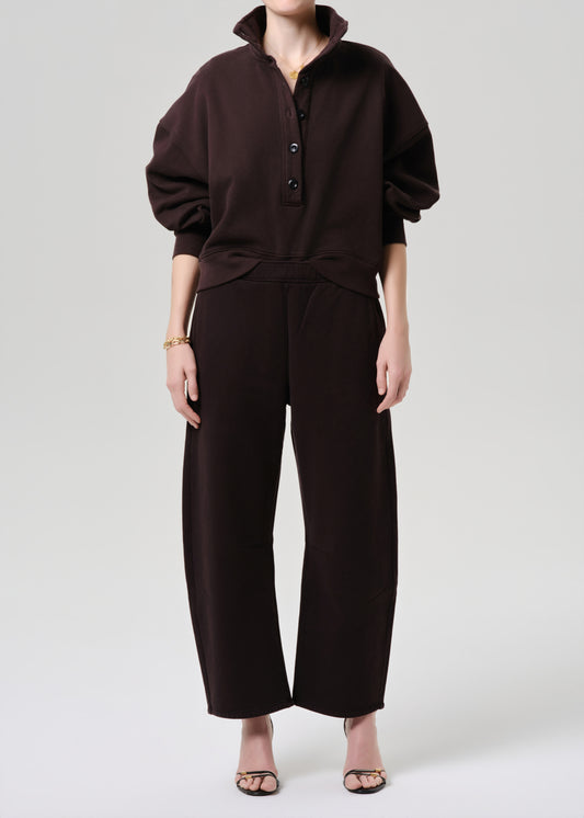 A model wearing a dark brown two-piece set consisting of a long-sleeve buttoned top and matching wide-leg cropped pants, paired with clear high-heeled sandals against a grey background.
