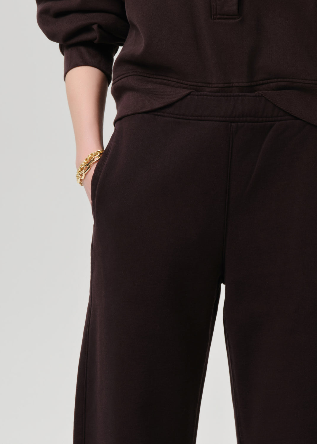 Close-up of a person wearing dark brown wide-leg sweatpants and a matching dark brown top, with one hand in a pocket and a gold bracelet visible, against a grey background.