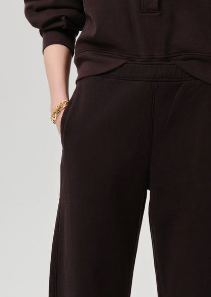 Close-up of a person wearing dark brown wide-leg sweatpants and a matching dark brown top, with one hand in a pocket and a gold bracelet visible, against a grey background.
