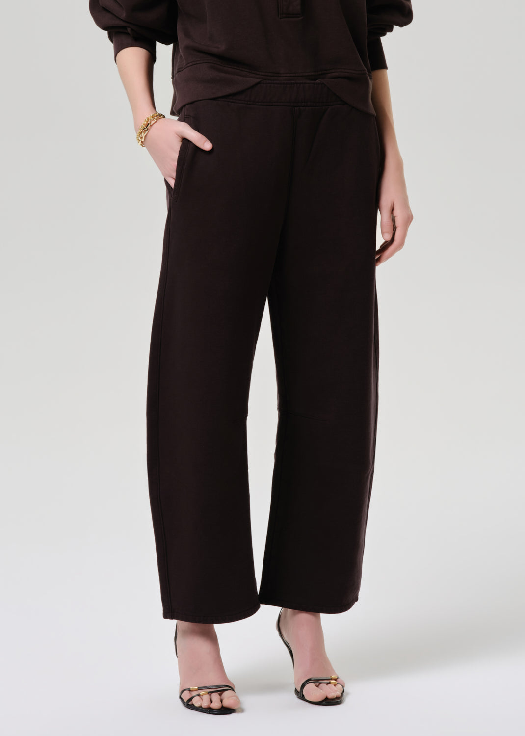 A model wearing dark brown wide-leg cropped trousers with side pockets, paired with a matching dark brown top and black strappy heels, shown from the waist down on a grey background.