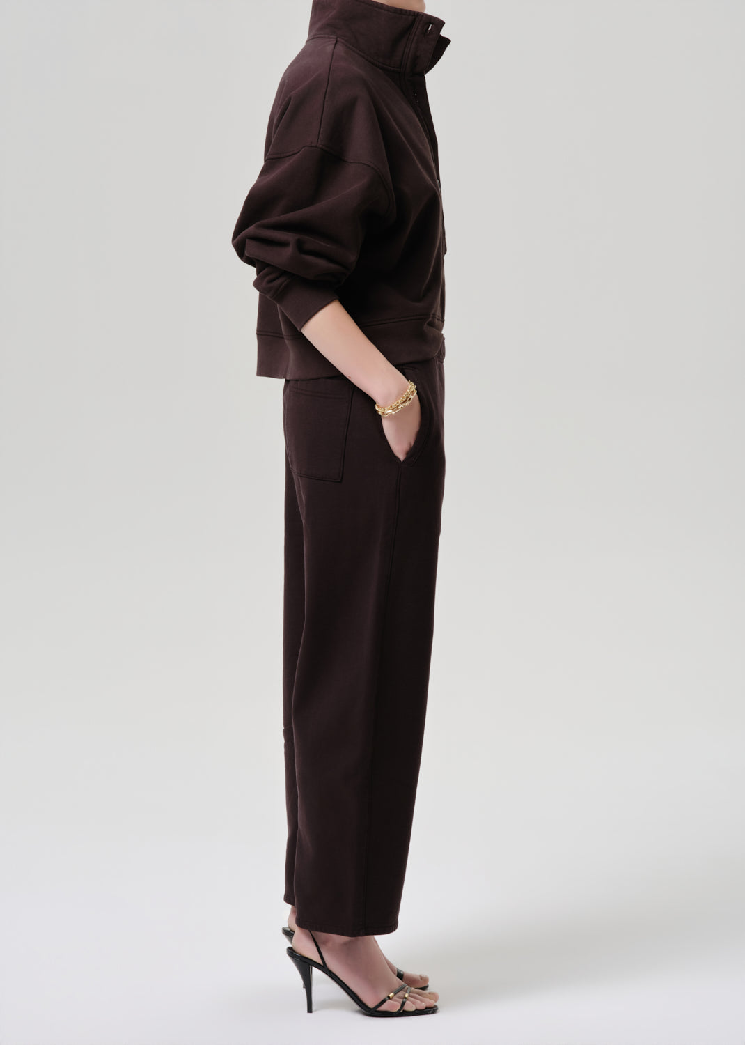 A model shown from the side wearing a dark brown cropped jacket and matching wide-leg trousers, paired with black high-heeled sandals against a grey background.