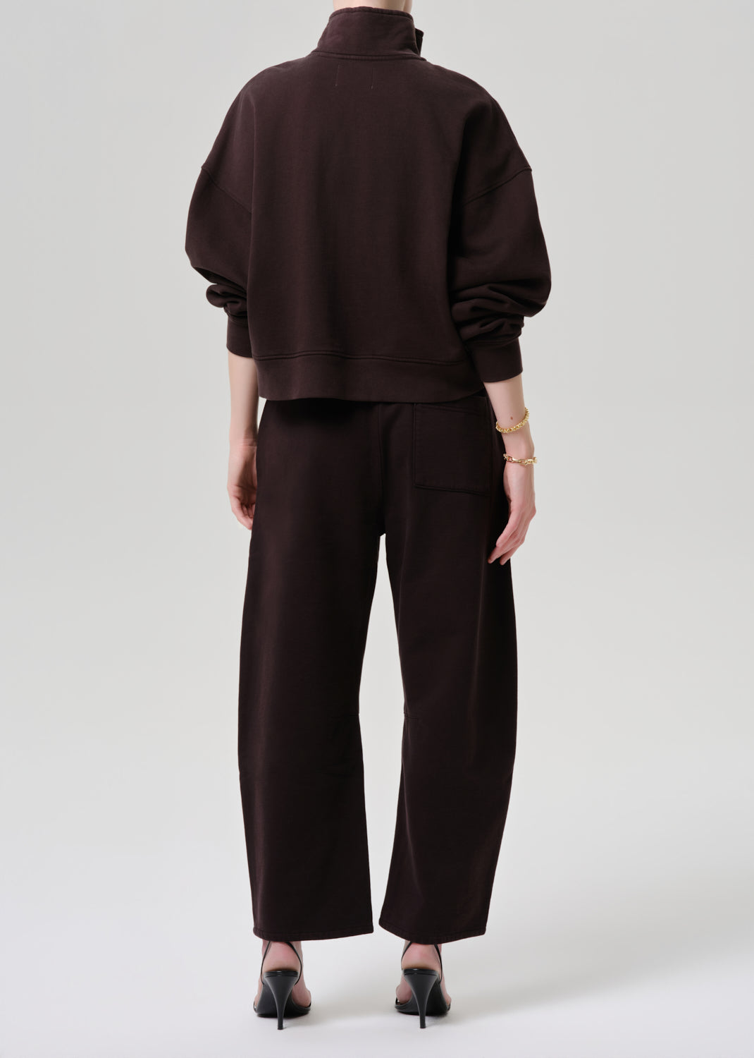 A model shown from the back wearing a dark brown cropped sweatshirt and matching wide-leg trousers, paired with black pointed-toe heels against a grey background.