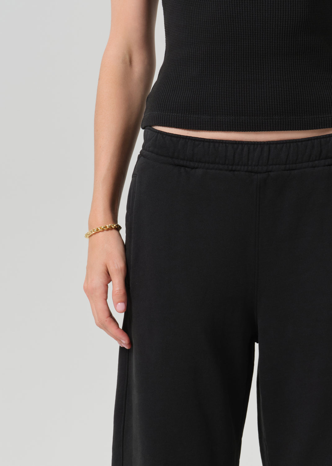 Close-up of a person wearing black wide-leg sweatpants and a black ribbed crop top, with a gold chain bracelet on one hand, against a grey background.