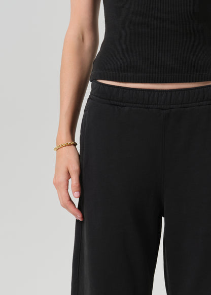 Close-up of a person wearing black wide-leg sweatpants and a black ribbed crop top, with a gold chain bracelet on one hand, against a grey background.