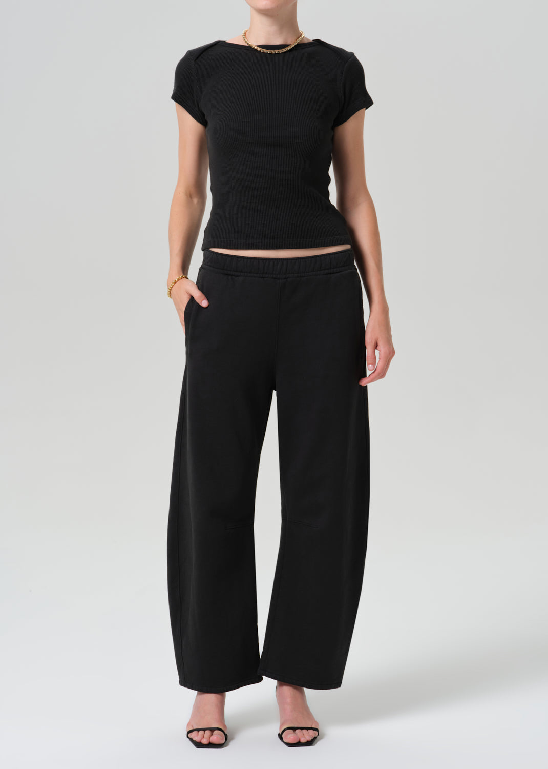 A model wearing a black short-sleeve cropped top and matching black wide-leg trousers, paired with black sandals, against a grey background.