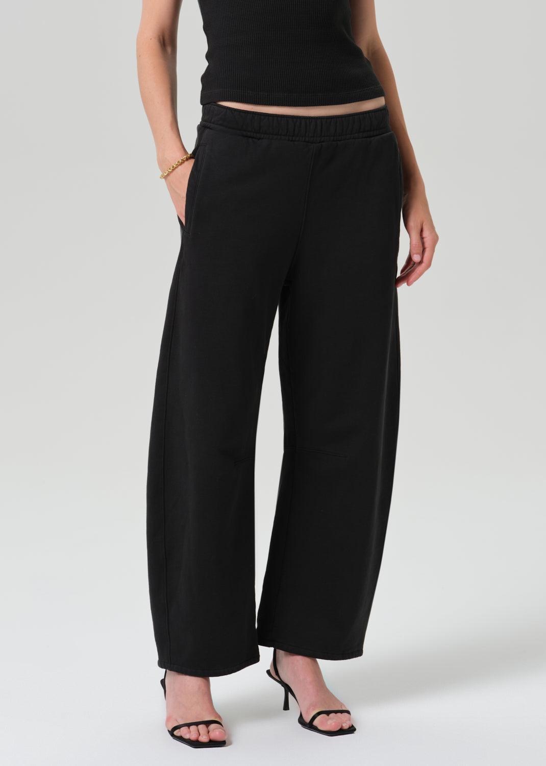 A model wearing black wide-leg cropped pants with an elastic waistband, paired with a black top and black heeled sandals, photographed against a grey background.