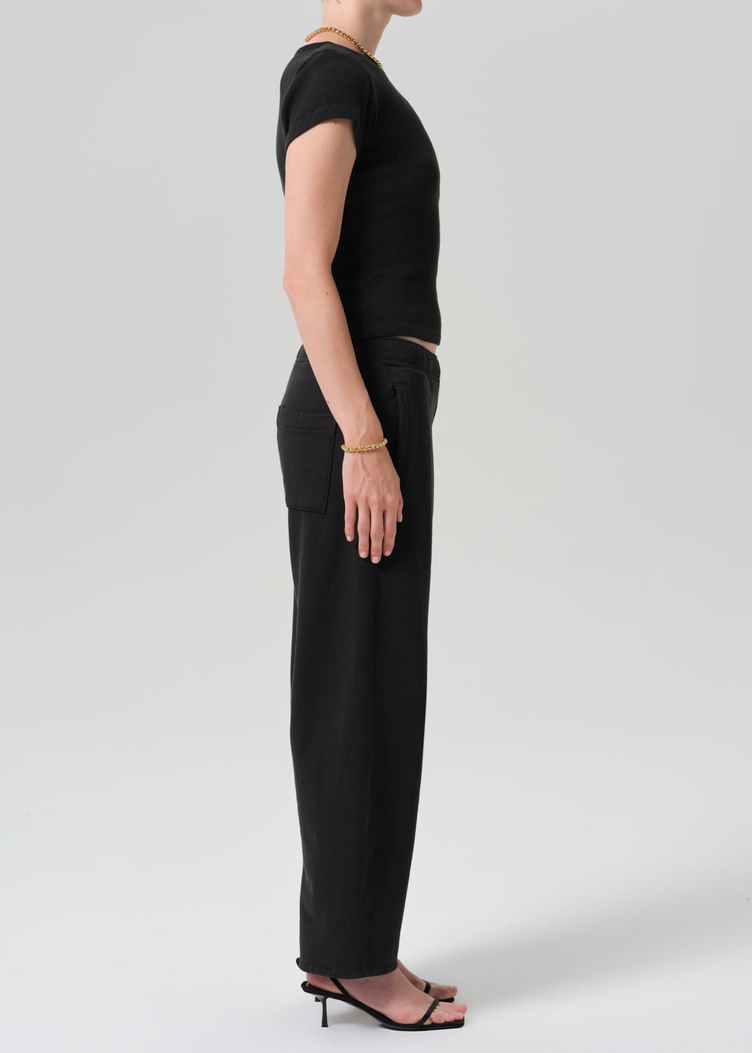 A model shown in profile wearing a black short-sleeve top and black wide-leg trousers, paired with black heeled sandals, against a grey background.
