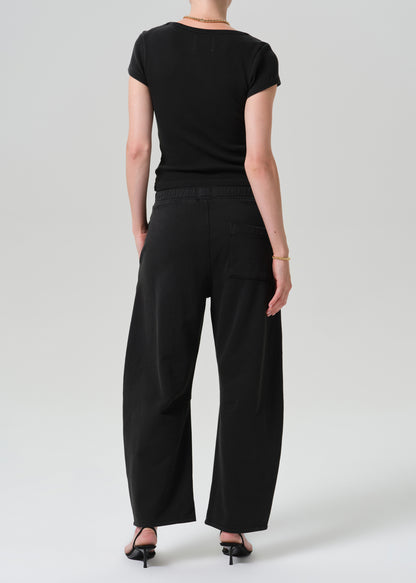 A model shown from the back wearing a black short-sleeve top and black wide-leg trousers, paired with black high-heeled sandals, against a grey  background.