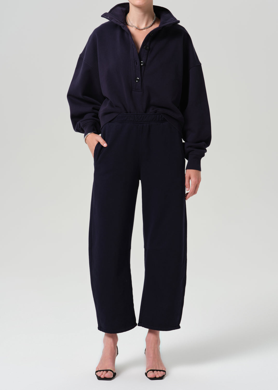 A model wearing a dark navy blue two-piece loungewear set consisting of a half-zip pullover hoodie and matching wide-leg cropped pants, styled with black heeled sandals against a grey background.