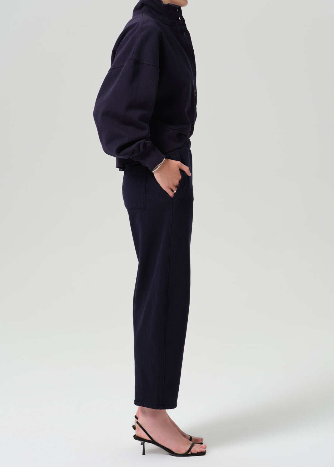 A model shown from the side wearing a dark navy blue two-piece set consisting of a cropped long-sleeve top and matching cropped trousers, paired with black heeled sandals against a grey background.