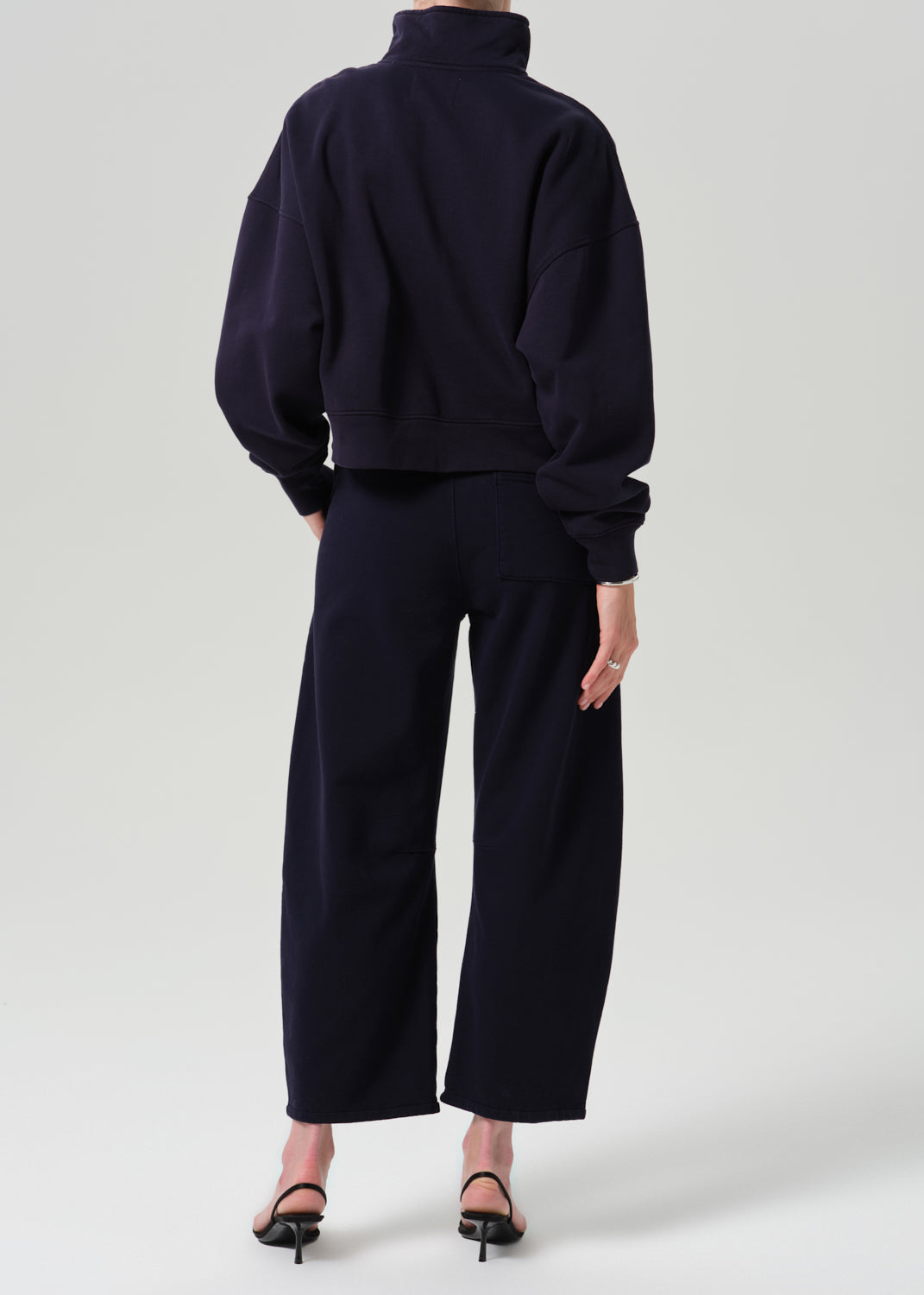 A model shown from the back wearing a dark navy blue cropped mock-neck sweatshirt and matching wide-leg cropped trousers, paired with black high-heeled sandals, against a grey background.