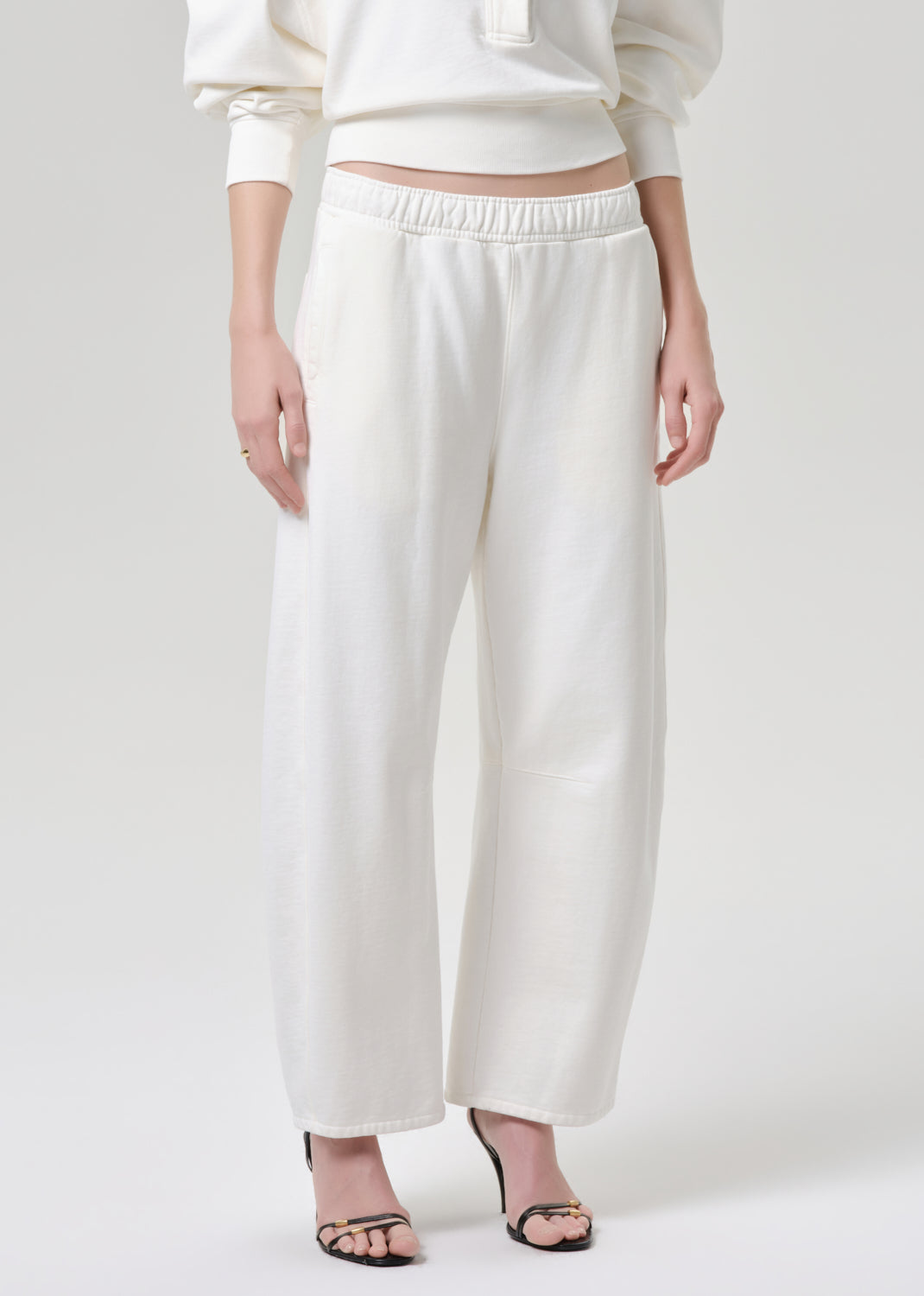 A model wearing off-white wide-leg cropped pants with an elastic waistband, paired with a matching cream sweatshirt and black heeled sandals, against a grey background.