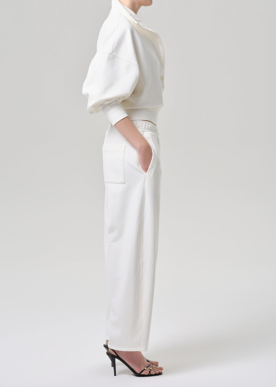 A model shown from the side wearing a white two-piece outfit consisting of a cropped top with voluminous puff sleeves and matching wide-leg trousers, paired with black high-heeled sandals against a grey background.