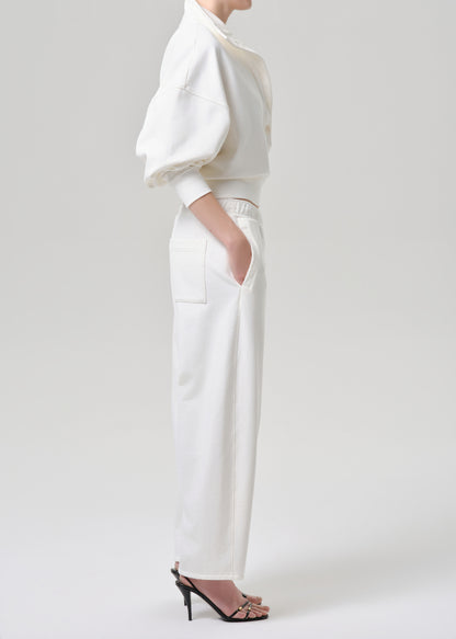 A model shown from the side wearing a white two-piece outfit consisting of a cropped top with voluminous puff sleeves and matching wide-leg trousers, paired with black high-heeled sandals against a grey background.