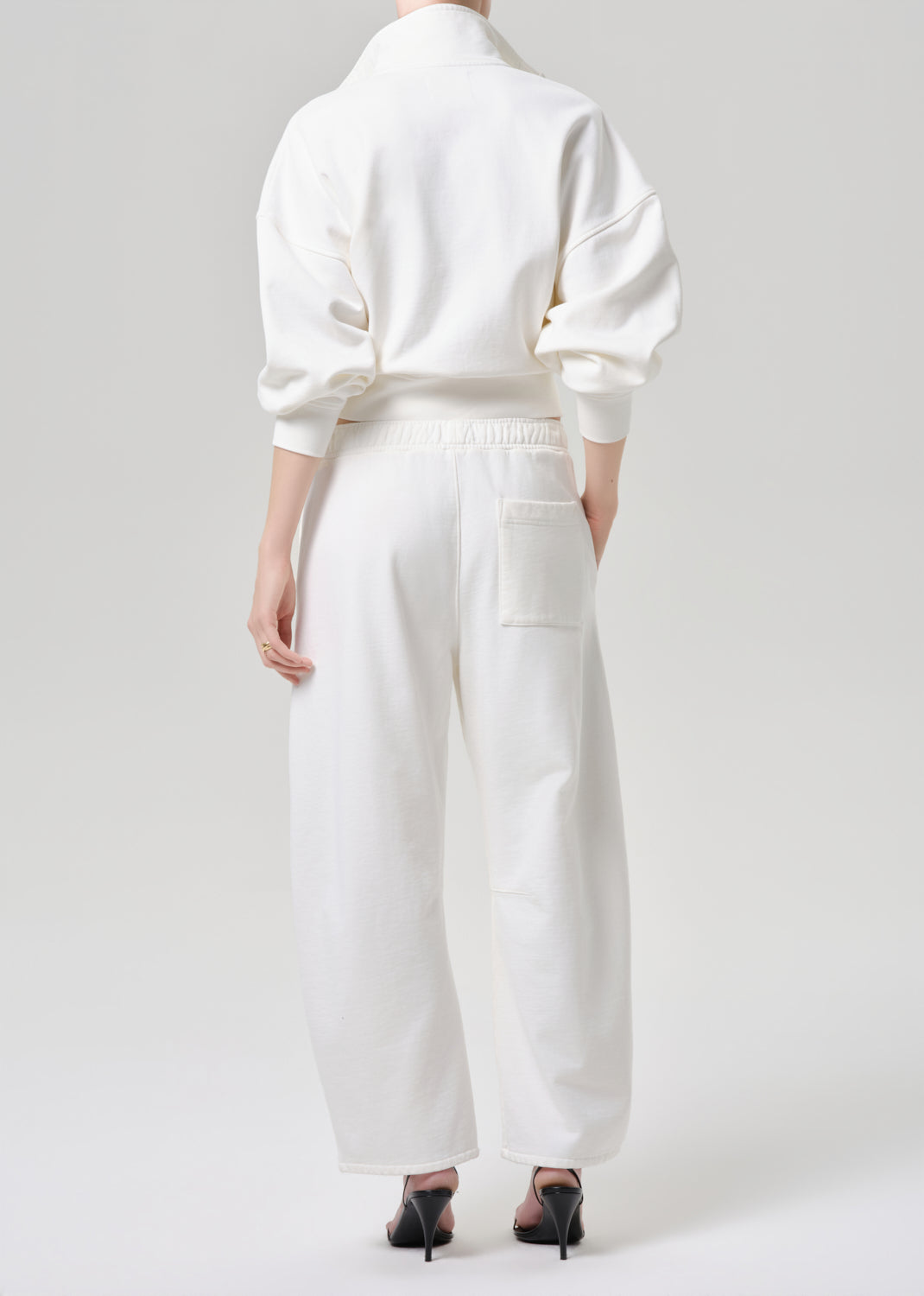 A model shown from the back wearing a white long-sleeve top and matching white wide-leg trousers with a back pocket, paired with black pointed-toe heels, against a grey background.