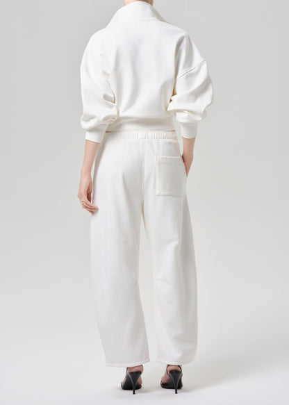 A model shown from the back wearing a white long-sleeve top and matching white wide-leg trousers with a back pocket, paired with black pointed-toe heels, against a grey background.