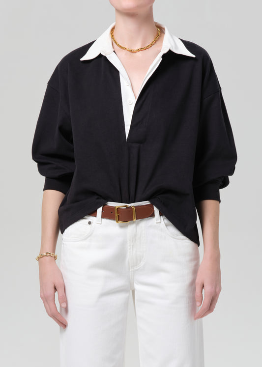 Person wearing a black rugby with a white collar, white pants, and a brown belt on a gray background