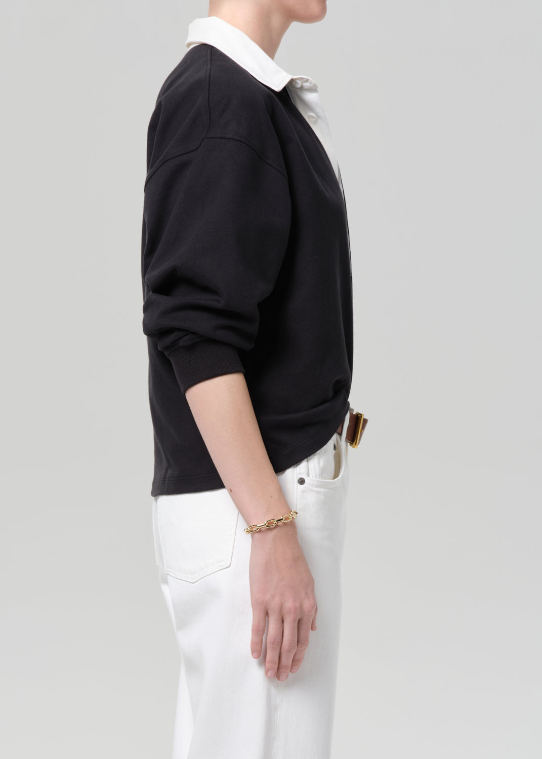Person wearing a black rugby with a white collar, white pants, and a brown belt on a gray background