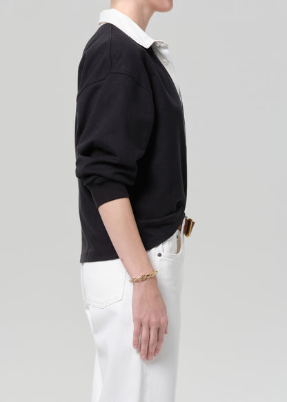 Person wearing a black rugby with a white collar, white pants, and a brown belt on a gray background
