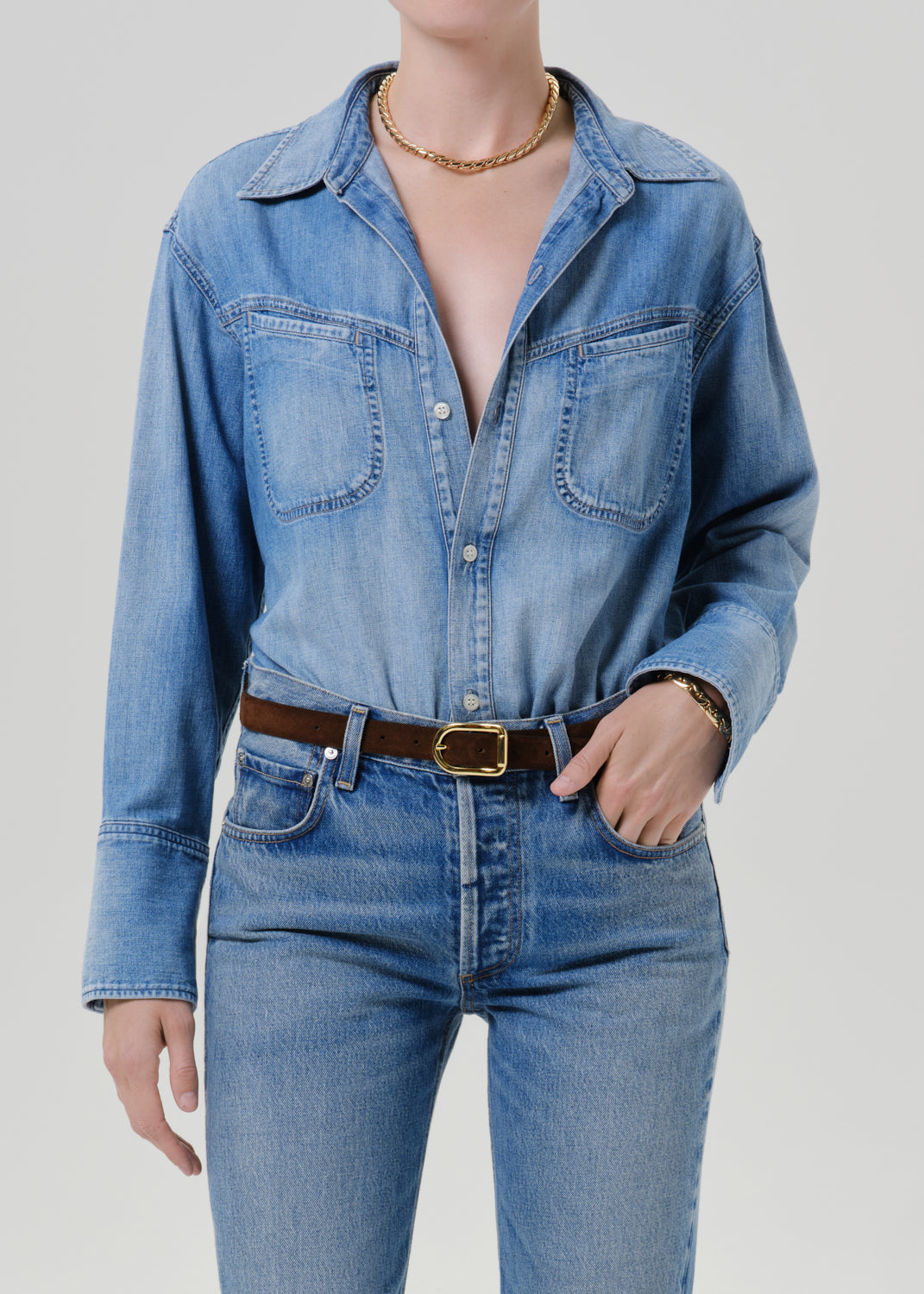A model wearing a light blue denim button-up shirt with chest pockets, tucked into matching blue jeans and styled with a brown leather belt with a gold buckle.