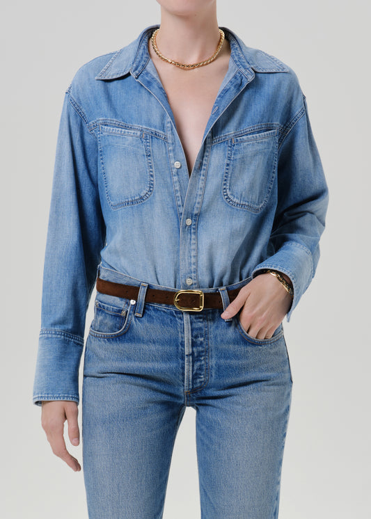 A model wearing a light blue denim button-up shirt with chest pockets, tucked into matching blue jeans and styled with a brown leather belt with a gold buckle.