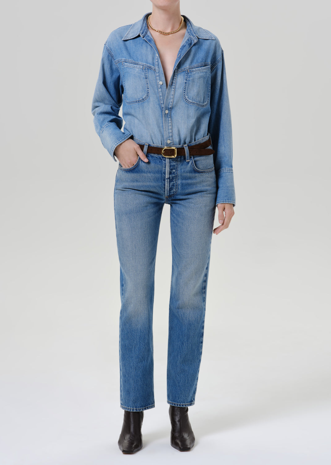 A model wearing a light blue denim shirt-jacket and matching straight-leg jeans, styled with a brown leather belt and black ankle boots against a grey background.