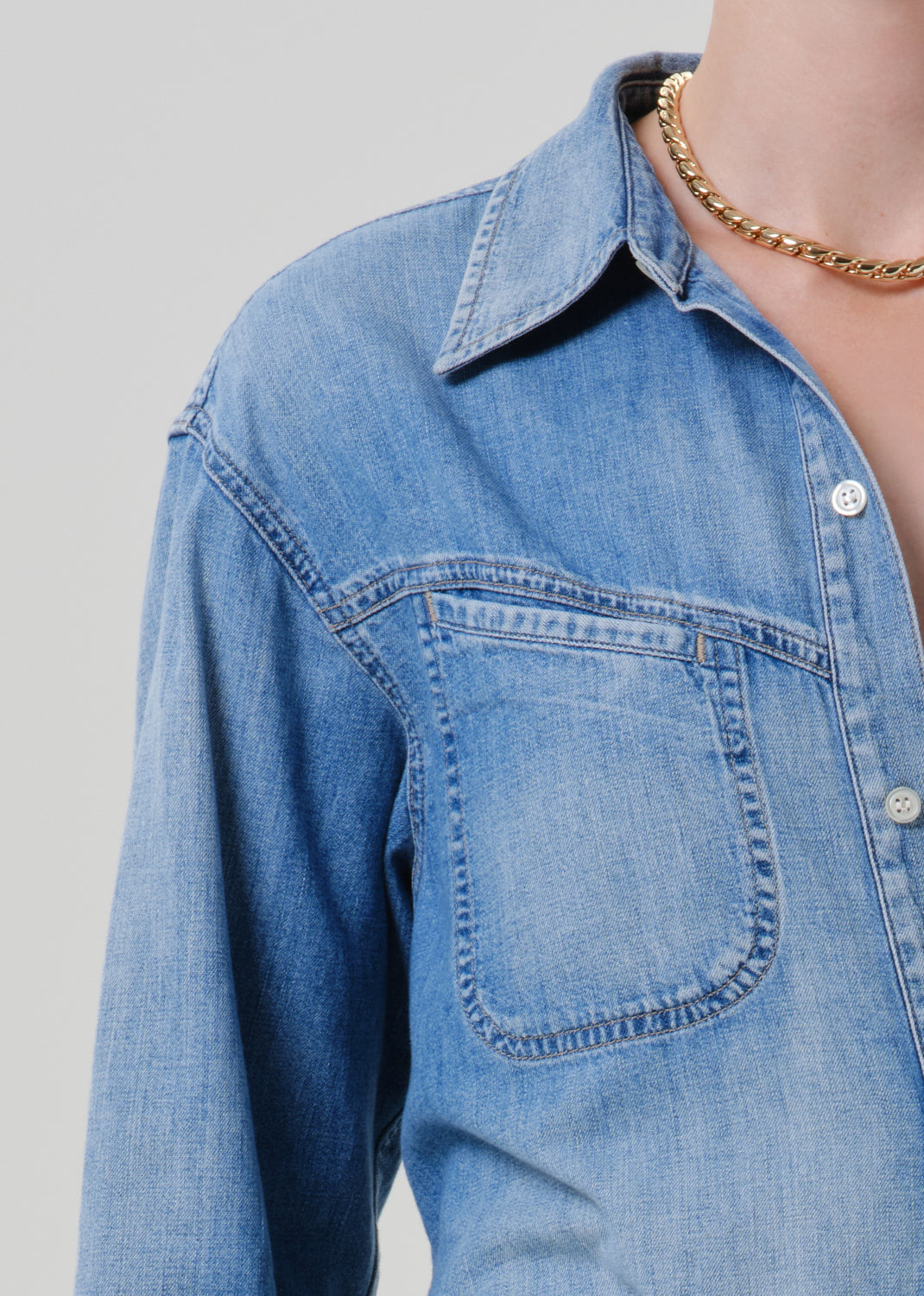 Close-up of a person wearing a light blue denim button-up shirt with a chest pocket and a gold chain necklace, against a grey background.