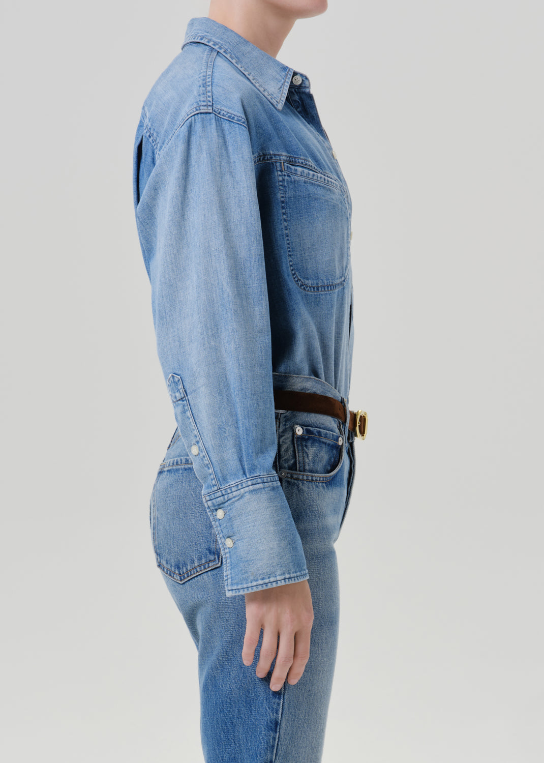 A side-profile view of a model wearing a light blue denim shirt-jumper with long sleeves, chest pockets, and buttoned cuffs, paired with matching blue jeans and a brown belt with a gold buckle, against a grey background.