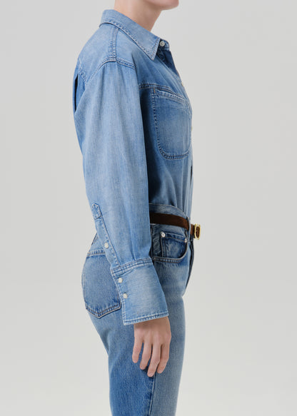 A side-profile view of a model wearing a light blue denim shirt-jumper with long sleeves, chest pockets, and buttoned cuffs, paired with matching blue jeans and a brown belt with a gold buckle, against a grey background.