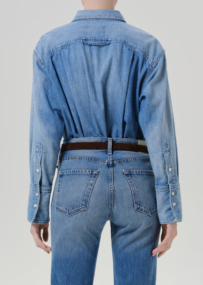 A model shown from the back wearing a light blue denim shirt tucked into matching blue jeans, accessorized with a dark brown belt, against a grey background.