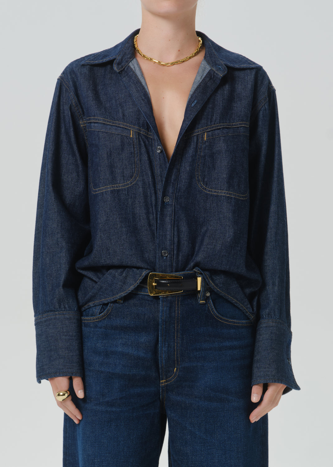 A model wearing a dark indigo denim long-sleeve button-up shirt with chest pockets, styled with a matching dark denim belt and dark blue jeans against a grey background.