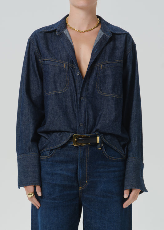 A model wearing a dark indigo denim long-sleeve button-up shirt with chest pockets, styled with a matching dark denim belt and dark blue jeans against a grey background.