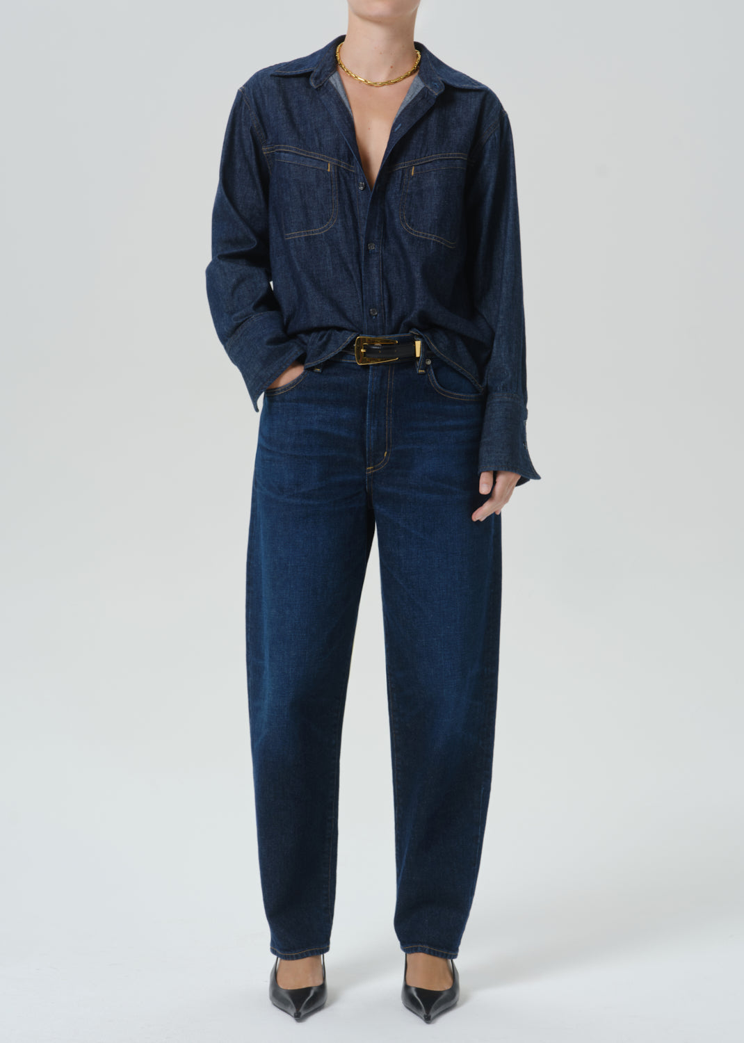 A model wearing a dark indigo denim shirt-jacket and matching straight-leg jeans, styled with a gold belt and black pointed-toe heels against a grey background.