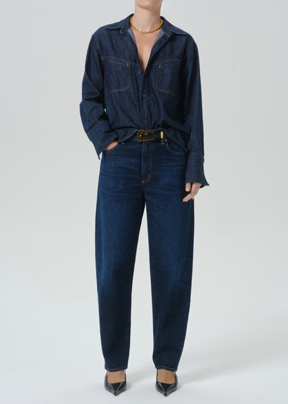 A model wearing a dark indigo denim shirt-jacket and matching straight-leg jeans, styled with a gold belt and black pointed-toe heels against a grey background.