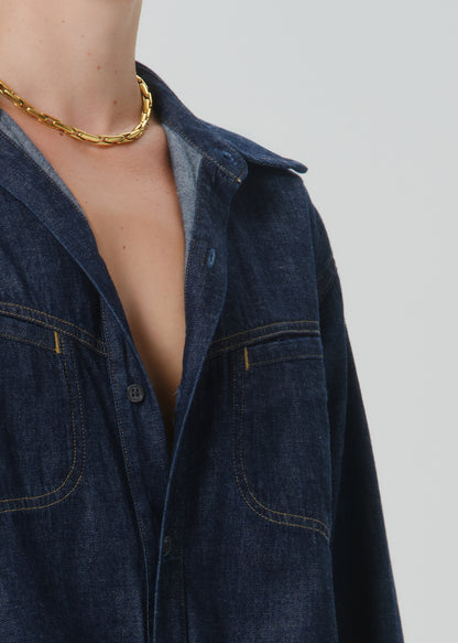 Close-up of a person wearing an oversized dark indigo denim shirt with contrast yellow stitching, a gold chain necklace, and a gray undershirt, set against a grey background.