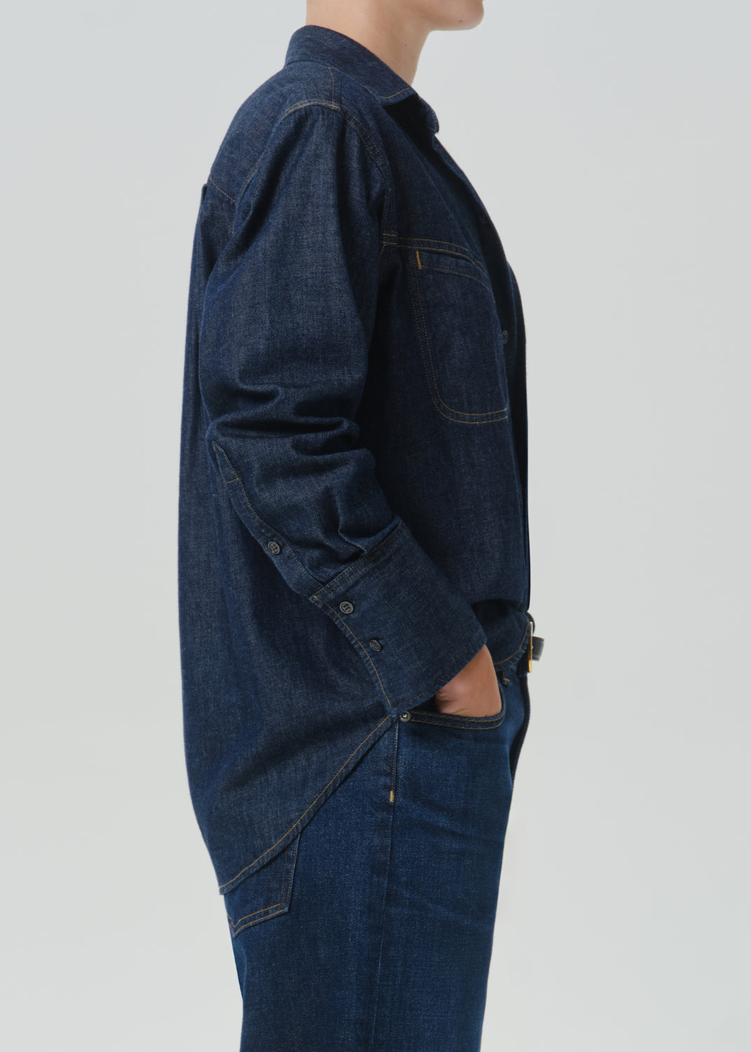 Side profile of a person wearing a dark indigo denim work jacket and matching dark denim jeans against a grey background.