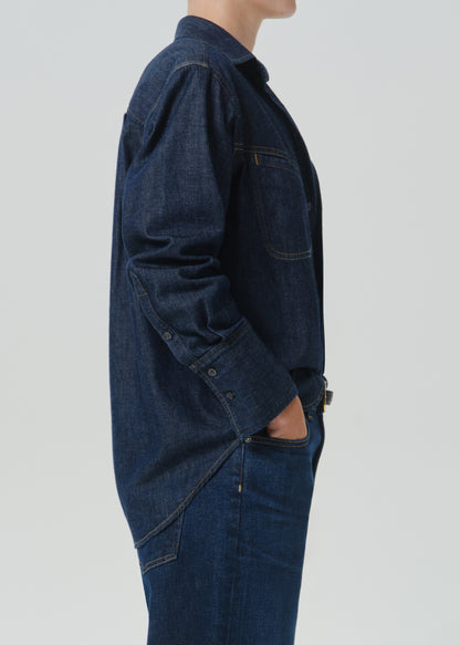 Side profile of a person wearing a dark indigo denim work jacket and matching dark denim jeans against a grey background.