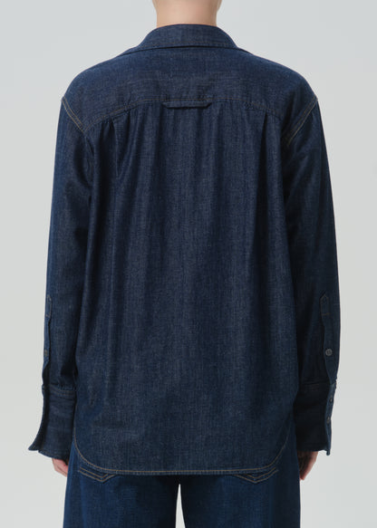A person shown from the back wearing a dark indigo denim long-sleeve button-up shirt and matching denim pants against a grey background.