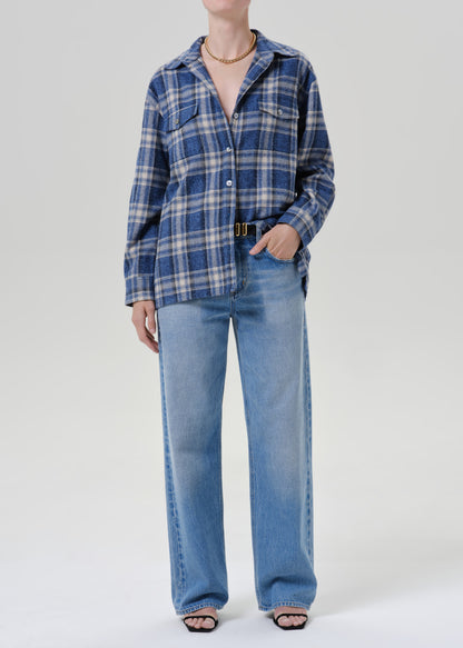 Person wearing a blue plaid button up shirt with light wash wide leg jeans and black sandals on a grey background