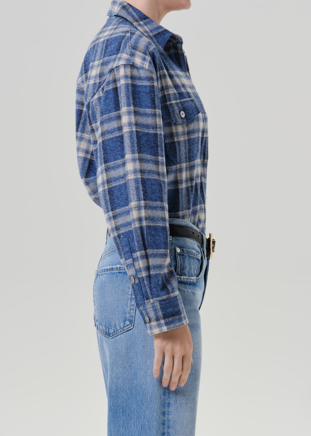 Person wearing a blue plaid button up shirt with light wash jeans viewed from the side on a grey background