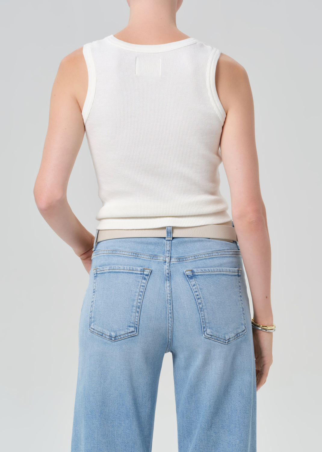 Person wearing a white tank top and light blue jeans with a belt on a gray background