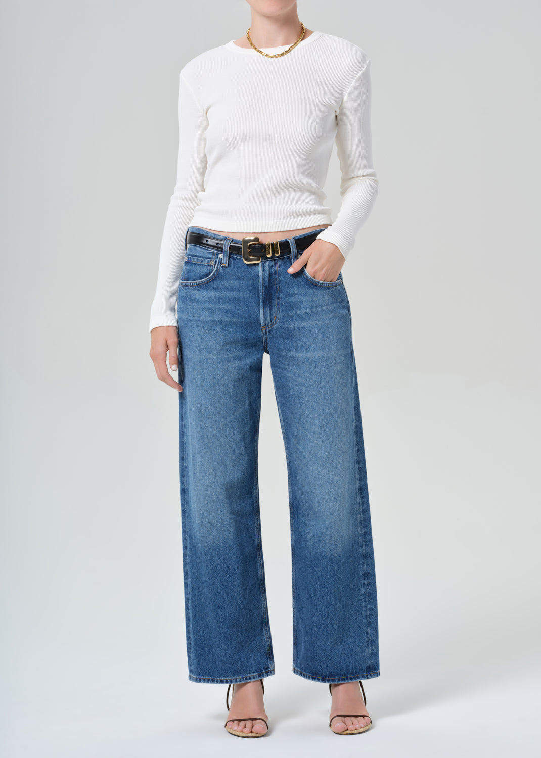 Person wearing a white long-sleeve top and blue jeans with a gold belt on a gray background