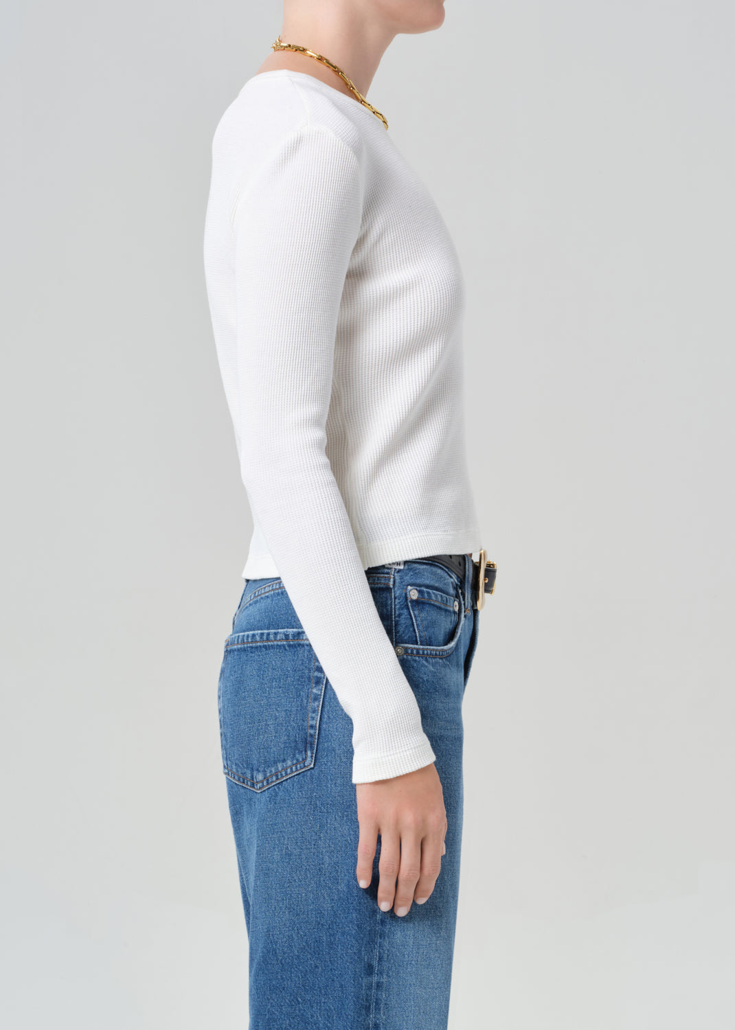 Person wearing a white long-sleeve top and blue jeans with a gold belt on a gray background