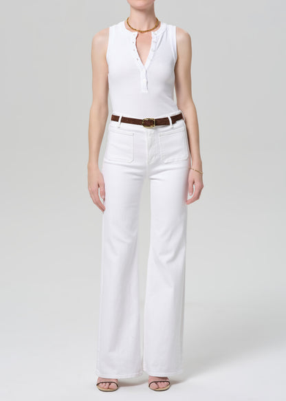 Person wearing a white sleeveless top and white jeans with a brown belt on a plain background