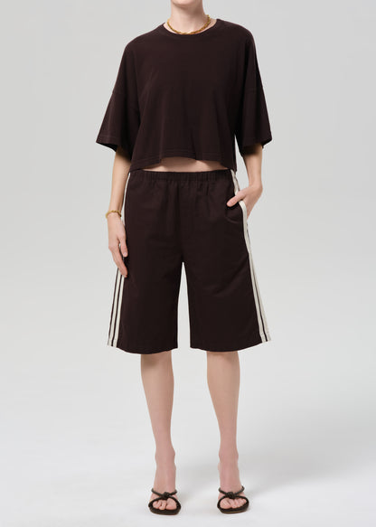 Person wearing a brown oversized t-shirt and brown shorts with white stripes on a light gray background