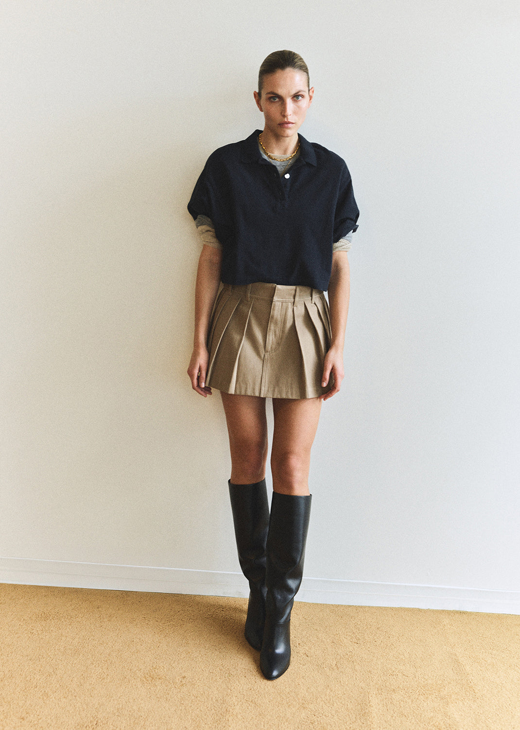 Person wearing a black top and beige skirt with black boots against a white wall.
