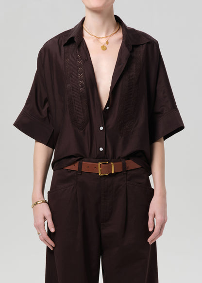 Person wearing a brown open-front shirt with a deep neckline and brown pants on a light gray background
