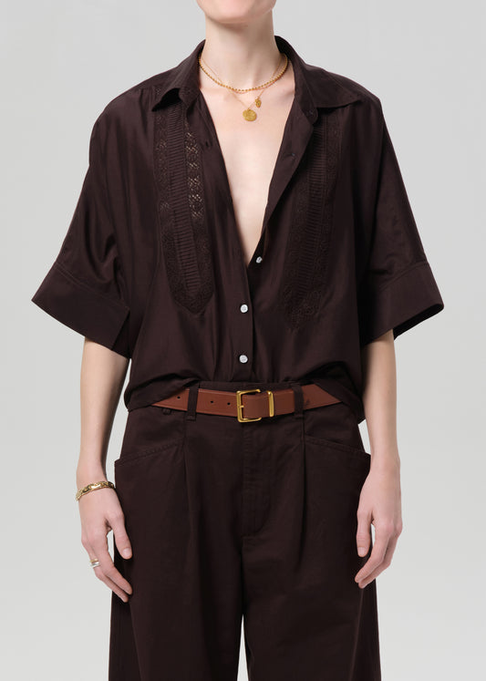 Person wearing a brown open-front shirt with a deep neckline and brown pants on a light gray background
